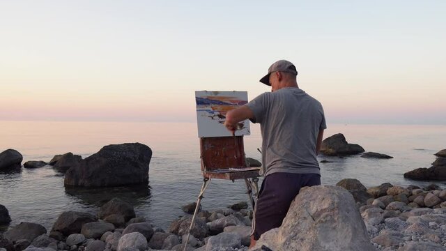 The artist man paints a seascape picture. Sea shore with rocks at dawn. Oil painting on canvas in nature. Plein air, landscape