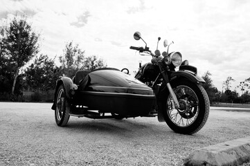 Black and white image of antique, vintage motorcycle with sidecar in open lot. - obrazy, fototapety, plakaty