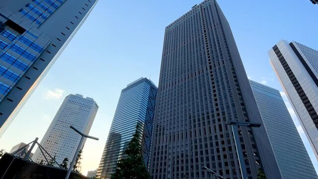SHINJUKU, TOKYO, JAPAN - JUNE 2021 : Exterior of tall office buildings in sunrise time. Wide low angle time lapse tracking shot, morning to day. Japanese urban metropolis and business concept shot.