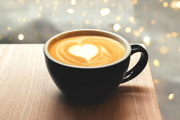 Black cup of coffee with heart shape latte art foam on wooden table in cafe with blurred lights bokeh