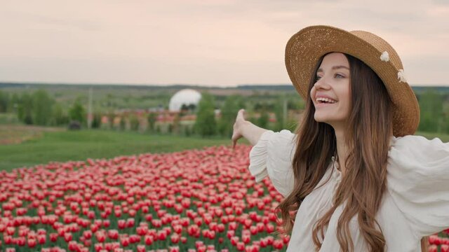Beautiful Smiling Girl on the Field with Tulips Enjoys Nature Breathes Freely. Standing at the Meadow with Colorful Flowers. Wears Straw Hat, having Romantic Mood, Long Hair. Cheerful Woman. Pretty.