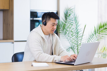 young asian man freelancer sitting at home at office desk with headset and talking online on video link via laptop in room or kitchen. The male indoors in casual clothes does the work and talks