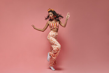 Wonderful funny girl with wavy dark hair and red lipstick in striped top and pink pineapple print pants dancing on isolated background..