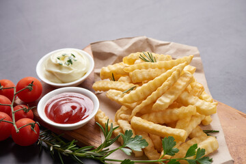Homemade baked potato fries with mayonnaise, tomato sauce and rosemary on wooden board. tasty french fries on cutting board, in brown paper on black stone table background, unhealthy food.