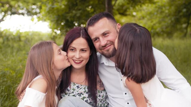 Two pretty sisters kissing their father and mother during summer picnic at green garden. Beautiful caucasian family relaxing together on fresh air. Love and happiness concept. Girls kissing Parents