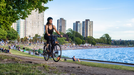 Fototapeta premium Adventurous White Caucasian Adult Woman riding a road bicycle in Stanley Park in a modern city. Sunny Summer Sunset. Downtown Vancouver, British Columbia, Canada.