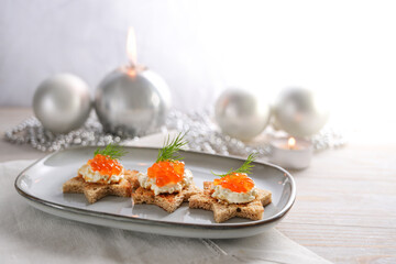 Festive red caviar canapes on toasted bread in star shape with cream and dill garnish on a gray plate, light wooden table with candles and silver Christmas decoration, copy space, selected focus