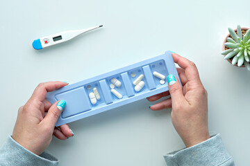 Patient hands hold box of pills. Plastic container with pills on off white table. Top view, flat lay with medications in mature caucasian female hands. Succulent plant and thermometer on table.