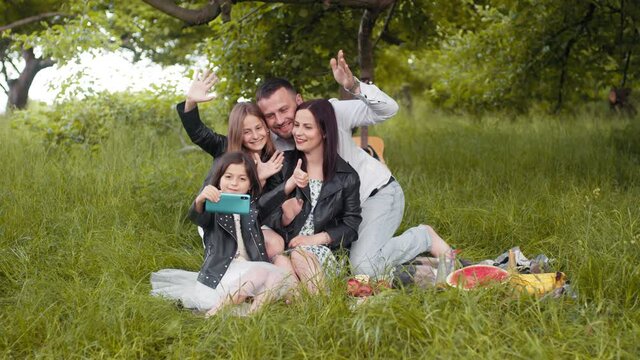 Cheerful parents and their cute daughters sitting together on green grass and using modern cell phone for taking self portrait. Concept of people, leisure and technology. Happy family taking selfie