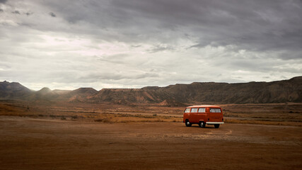 orange camper van parked in the desert for the night, red sand and mountains in the background with a cloudy sky