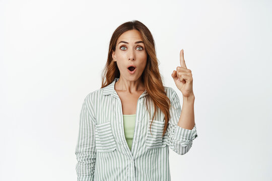 Image of surprised middle aged woman, businesswoman pointing finger up with WOW face, checking out smth awesome, impressive big sale, standing over white background