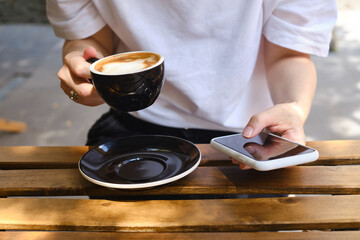 Girl drinks coffee from a black cup and uses a smartphone. A woman is holding a mobile phone, sending a text message or using an application on her mobile phone. Drink cappuccino or latte in a cafe
