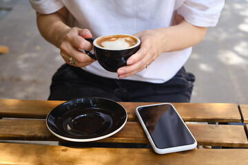 Fototapeta na wymiar Girl drinks coffee from a black cup and uses a smartphone. A woman is holding a mobile phone, sending a text message or using an application on her mobile phone. Drink cappuccino or latte in a cafe