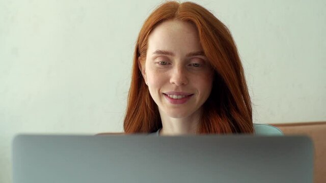 Close-up front view of smiling redhead young woman with no makeup working typing on laptop computer sitting at table at home office. Portrait of cheerful attractive lady using laptop looking at screen