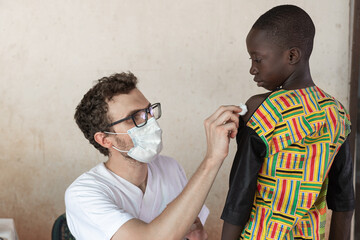 Doctor with glasses cleaning shoulder before performing injection on African little boy.