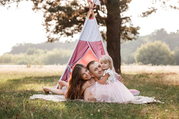 Mom, dad and little daughter are sitting next to wigwam decoration in the park. Family spending time outdoor in summer, having fun together. father's, mother's and baby's day © Andriy Medvediuk