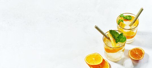 Close up of two glassas of summer trendy sparkling cocktail with gin and cold tonic, sliced orange, mint, ice cubes with bamboo straw on white background with copy space. Wide banner.