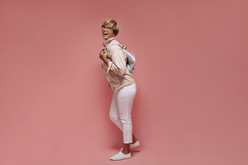 Full length photo of woman with short hair and glasses in skinny light pants, white sneakers and jacket laughing and holding bag on pink backdrop..