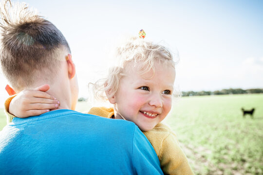 Little Sister Smiles. Girl Looks Out From Behind Her Brother's Shoulder. Teenager Carries A Child In Arms Across The Field.