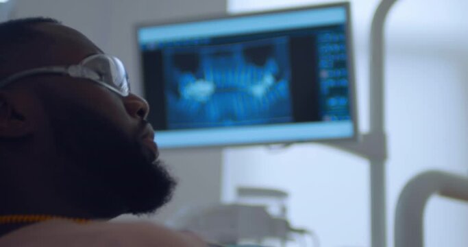 A close-up monitor screen with a picture of teeth computed tomography scan and an African American male patient in disposable eyeglasses is looking at something next to him.