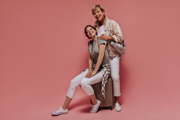 Trendy young girl in sneakers, light trousers and t-shirt smiling, sitting on suitcase and posing with short haired woman on isolated backdrop..