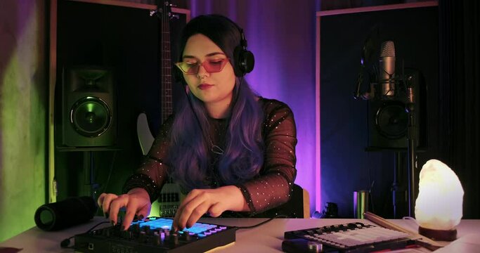 Medium shot of female LGBTQ non-binary producer writing music on push midi controller synthesizer with long colored hair in studio. Dj music session beautiful singer composer, festival moody vibe