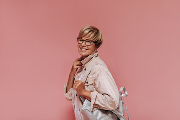 Smiling woman with stylish blonde hairstyle, glasses and grey bag in beige modern jacket looking into camera on pink backdrop..