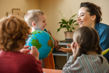happy boy holding globe near cheerful teacher an kids on blurred foerground