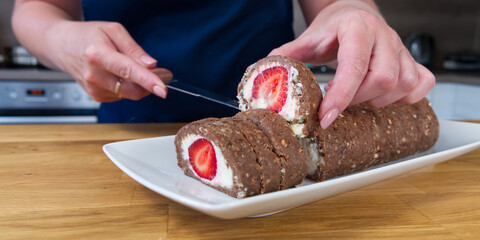 Woman's hands cutting a piece of Chocolate roll with curd cream and strawberries inside. Strawberry dessert on the home wooden table. close up