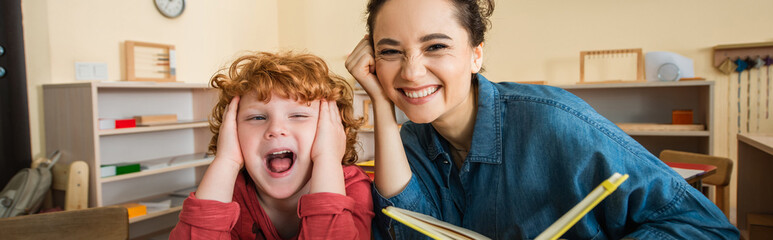cheerful teacher holding book near redhead astonished boy, banner