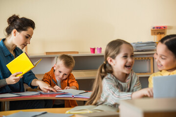 teacher pointing at notebook near writing boy while interracial girls laughing on blurred foreground
