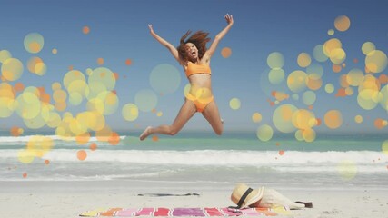 Animation of african american woman jumping for joy at beach over yellow spots - Powered by Adobe