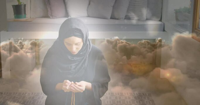 Animation of asian woman in hijab praying at home over clouds
