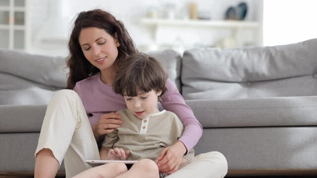 Young single mother or baby sitter relax with small boy and digital tablet: mom and kid sitting cuddling with touchscreen computer in hands smiling watch funny video or cartoon online at home together