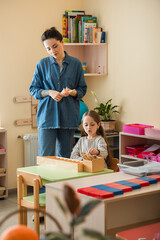 Obraz premium young teacher standing near girl playing educational game in montessori school