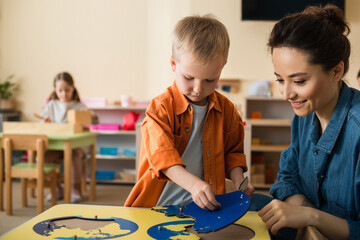 boy combining earth map puzzle near smiling teacher and girl on blurred background