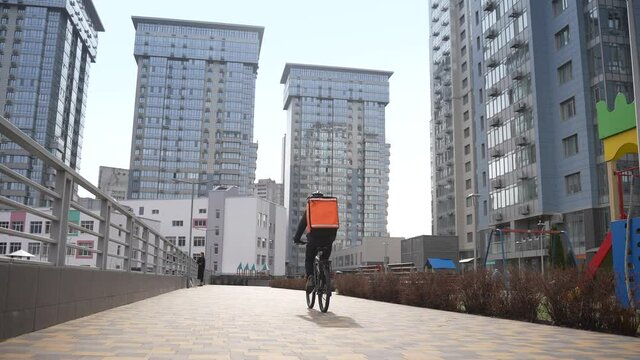 Back view of courier cyclist carrying thermal backpack while riding bicycle past urban playground surrounded by high-rise buildings. Delivery guy with orange insulated bag during work around city