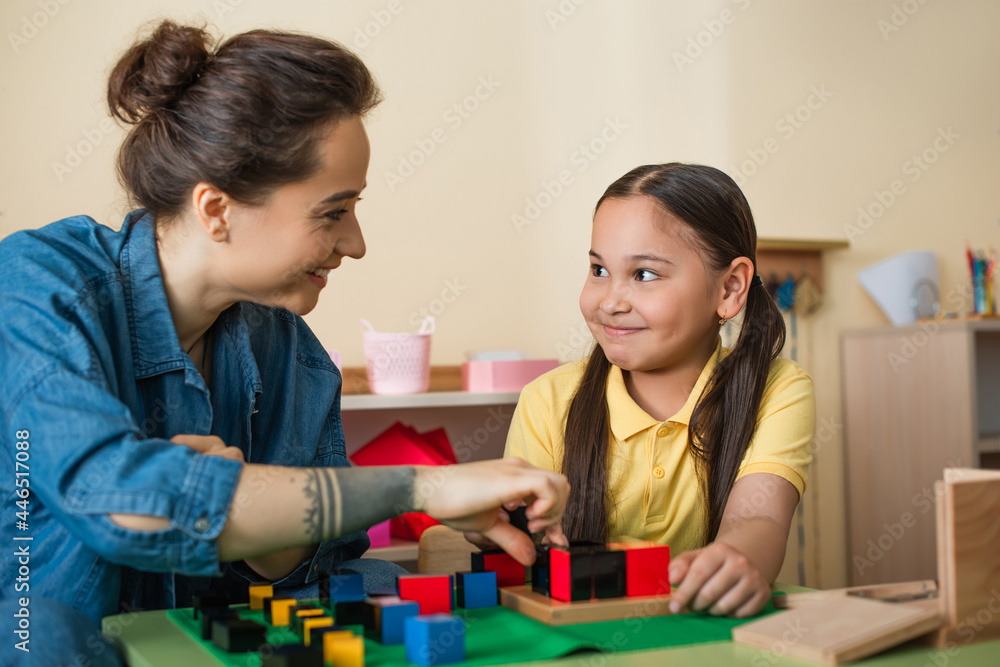 Wall mural happy asian girl and young teacher looking at other while playing with wooden cubes