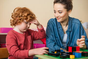 teacher smiling near thoughtful boy and multicolored cubes in montessori school
