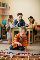 Fototapeta premium boy on floor pointing at wooden letters near teacher and interracial kids on blurred background