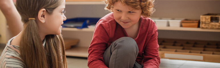 redhead boy smiling while looking at girl in montessori school, banner
