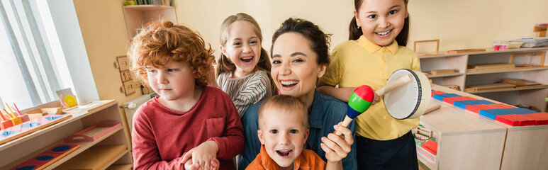 cheerful teacher looking at camera while playing music with interracial kids, banner