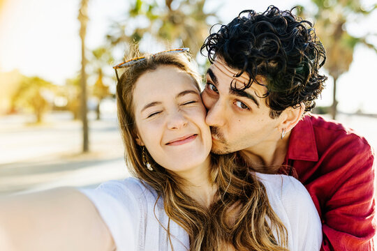 Young happy man kissing cheerful hispanic woman with closed eyes while taking selfie on smartphone in the beach - Funny young couple having fun during summer holidays - Relationship and friendship