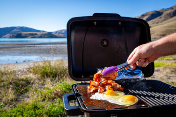 A man cooks bacon and eggs on a portable bbq beside the seaside on a sunny winter day travelling in...