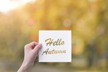 Hand holding paper with Hello Autumn text over blur nature background. Autumn concept.