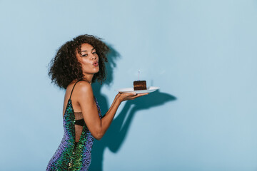 Stylish girl with curly brunette hairstyle in green modern dress holding piece of cake with candle on isolated background..