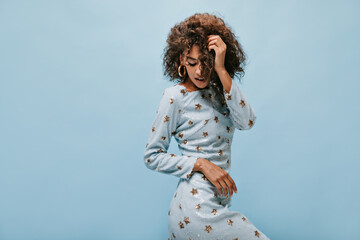 Attractive lady with brunette curly hairstyle in trendy shiny dress and round modern earrings posing on blue background..