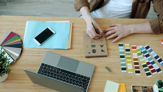 High Angle View Of Male Hands Designer Sketching Logo Symbols And Typing With Laptop Working In Office At Desk. Technology And Creative Work Concept.