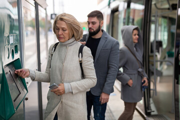 Focused mature woman in light overcoat buying ticket for public transport in street vending machine in autumn day
