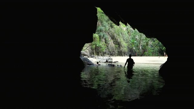 Hand held shot of male snorkeler entering emerald cave in Koh Muk Thailand. A silhouette exits the ocean water and walks through a dark cave towards the white sand beach with karst mountain background
